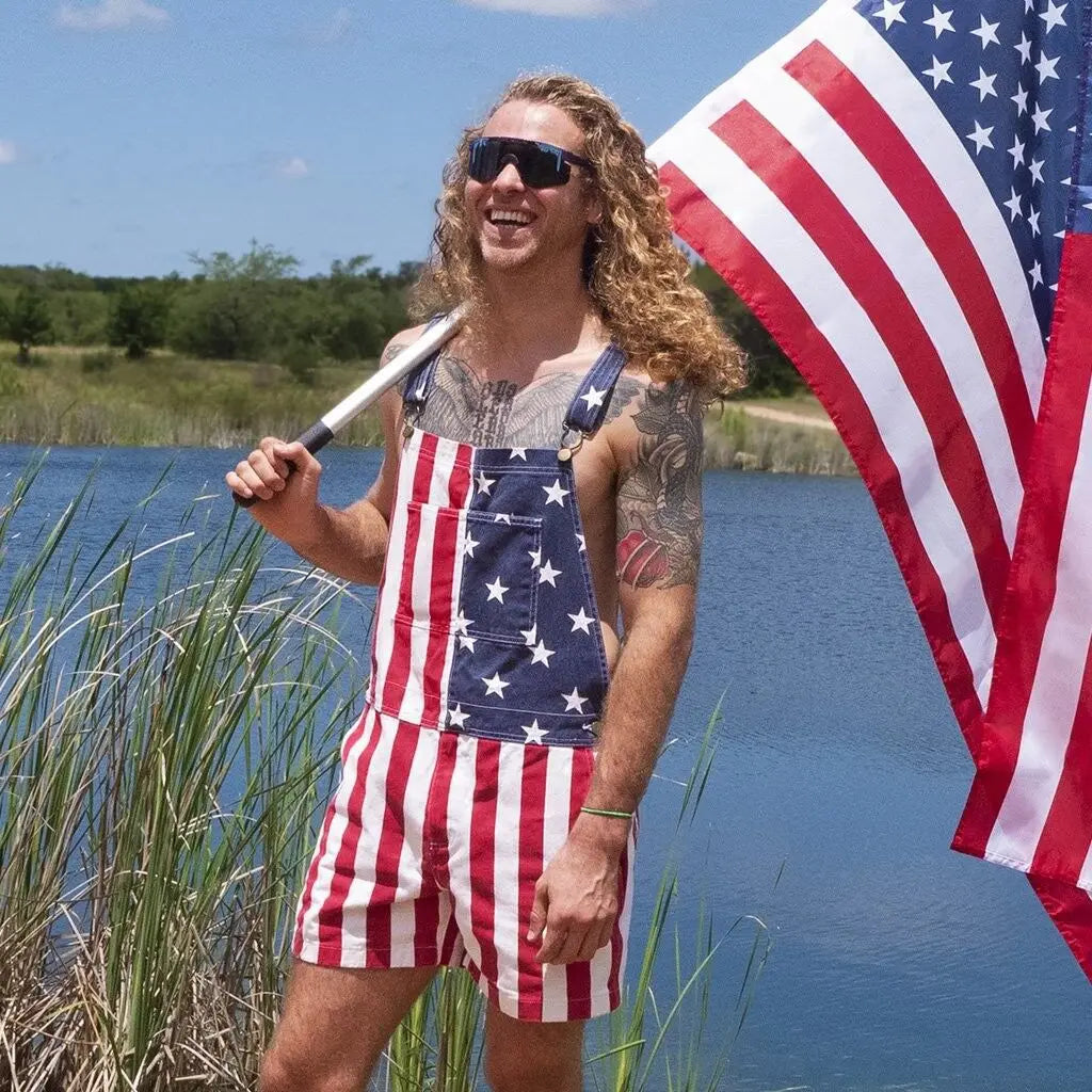 A Long haired man wearing american flag overalls while holding the USA flag with a lake behind him